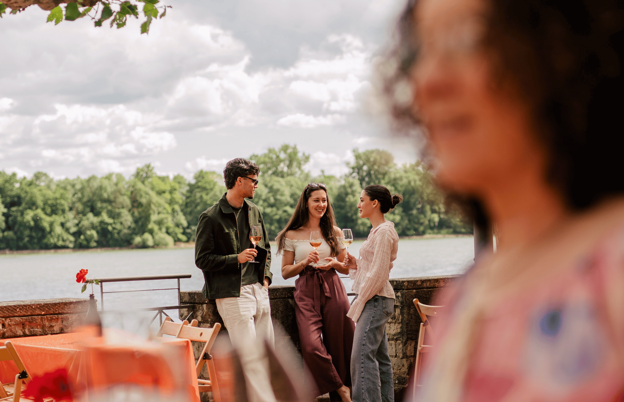 Ein Mann und zwei junge Frauen die ein Glas Sekt in der Hand haben, sich unterhalten und im Sektgarten mit Blick auf den Rhein stehen